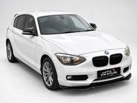 Load image into Gallery viewer, A white BMW 1 Series hatchback car shown from a front three-quarter angle on a light gray studio background. The car features a black front lip spoiler, black kidney grilles, and a front license plate frame with the text 'STM STYLING'.
