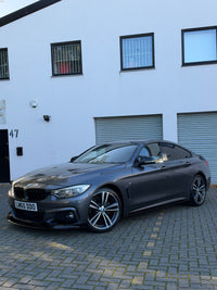 Load image into Gallery viewer, A dark gray BMW 4 Series coupe parked on a gray paved driveway in front of a white building with garage doors and windows.
