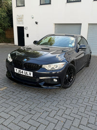 Load image into Gallery viewer, A black BMW 4 Series coupe with a front splitter and black wheels, parked on a gray paved driveway in front of a white building.
